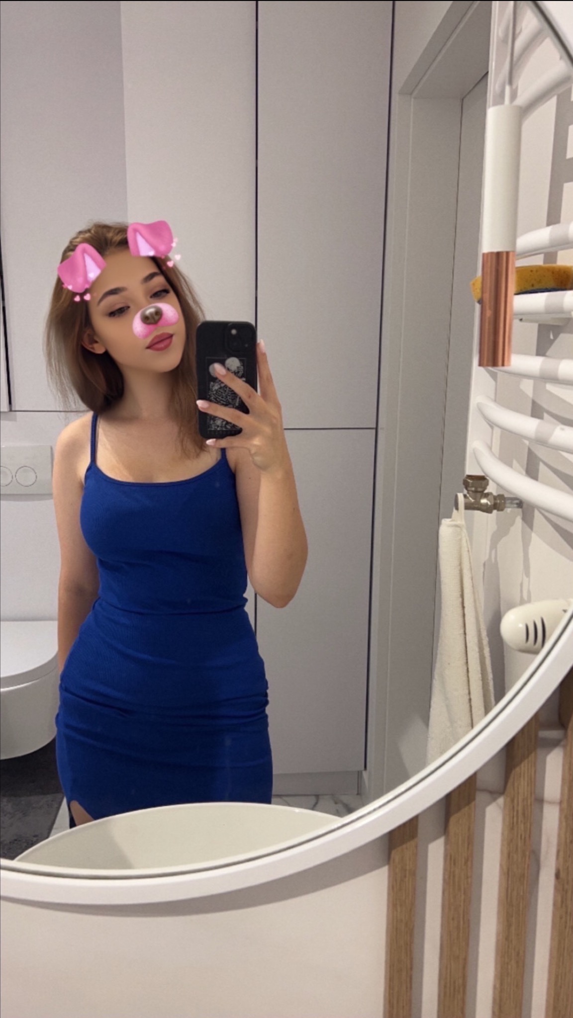 Młoda kobieta robi selfie w łazience z filtrem z uszami i nosem pieska, widoczna umywalka, grzejnik drabinkowy i białe szafki.