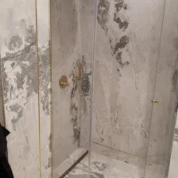 Kabina prysznicowa z marmurowymi płytkami w odcieniach szarości i bieli, złota armatura i listwy wykończeniowe, widoczny odpływ liniowy.