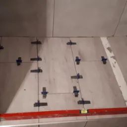 Układanie płytek imitujących drewno na podłodze łazienki, widoczne krzyżyki dystansowe i poziomica, ściany częściowo wykończone szarą glazurą.