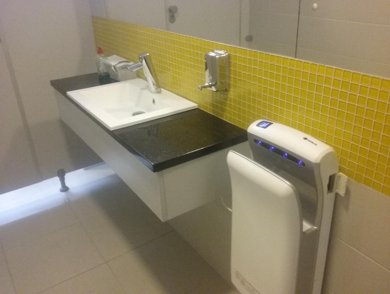Umywalka z czarnym blatem i baterią, żółta mozaika na ścianie, podajnik mydła w płynie i suszarka do rąk w publicznej toalecie.