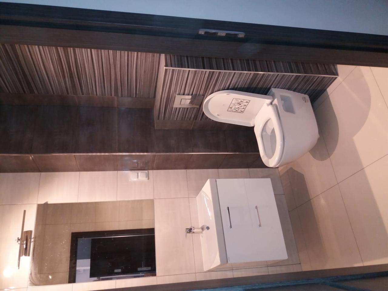 Ukończona łazienka w Gdańsku z toaletą z deską sedesową z instrukcjami, umywalką z szafką i dużym lustrem, wykończona płytkami imitującymi drewno.