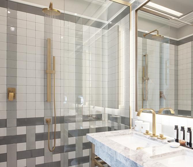Nowoczesna łazienka z kabiną prysznicową typu walk-in, wykończona białymi i szarymi kwadratowymi płytkami oraz złotą armaturą, woda leje się z deszczownicy.
