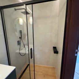 Elegancka, nowoczesna łazienka w minimalistycznym stylu. Jasne płytki i drewniana podłoga nadają wnętrzu ciepła, a okrągłe lustro w złotej ramie i subtelne oświetlenie LED podkreślają wyjątkowy charakter przestrzeni.