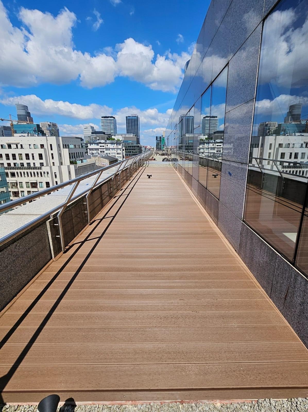 Długi taras na dachu budynku z deskami w odcieniach brązu, widok na panoramę miasta odbijającą się w szklanej fasadzie, barierka ze stali nierdzewnej.