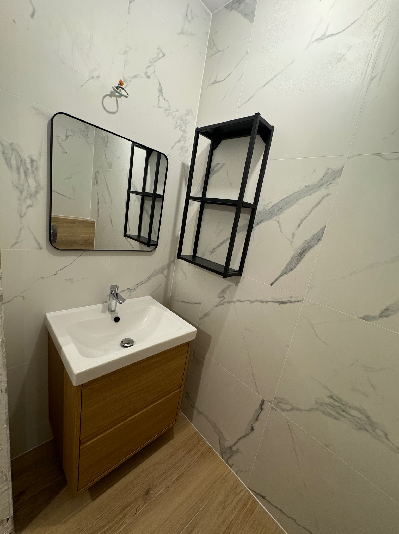 Nowoczesna łazienka z umywalką nablatową na drewnianej szafce, czarnym lustrem i półką na tle białych płytek imitujących marmur, widoczny drewniany parkiet.