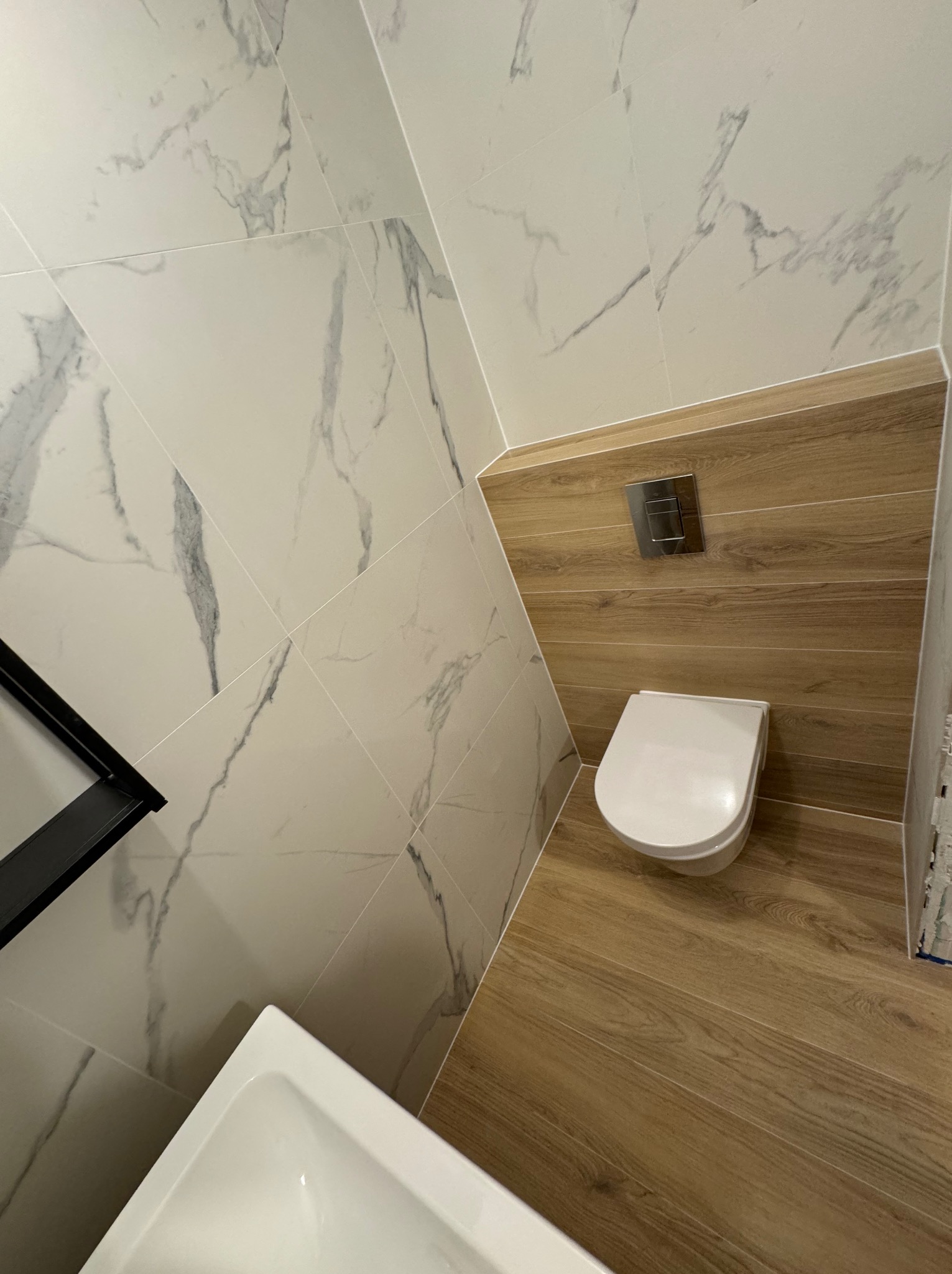 Nowoczesna toaleta w Piasecznie z drewnopodobnymi płytkami na podłodze i ścianie za muszlą klozetową, kontrastująca z marmurowymi płytkami na pozostałych ścianach, widoczna umywalka i czarna półka.