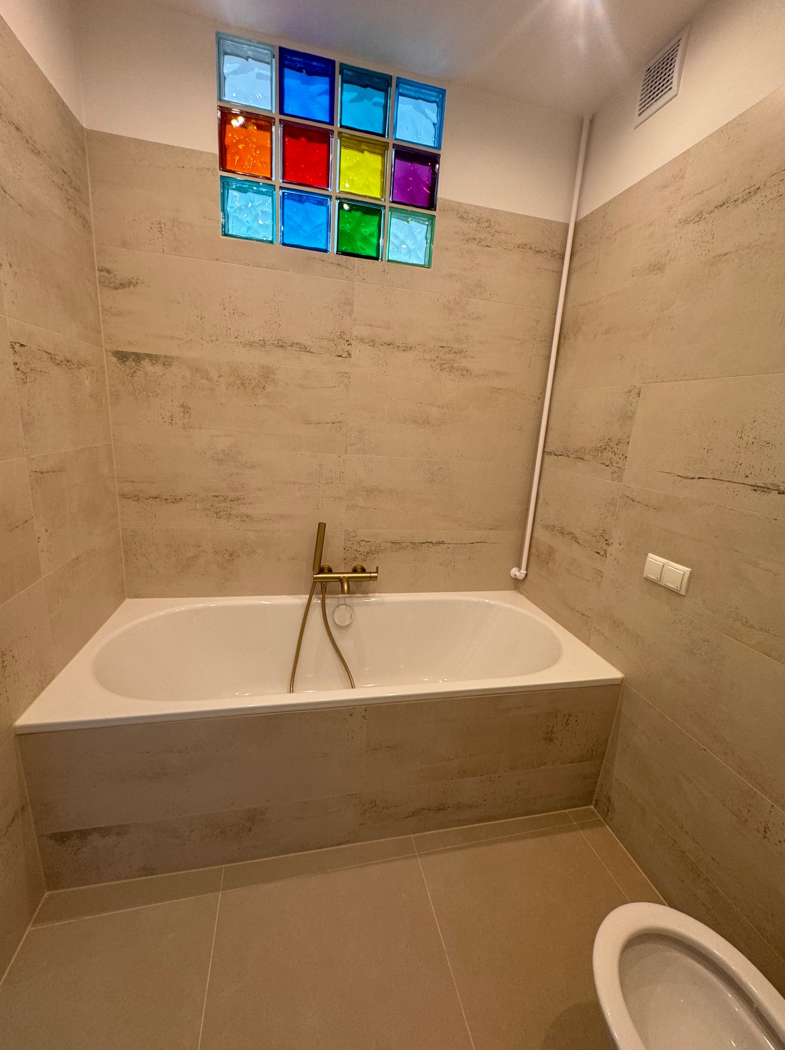 Jasna łazienka z beżowymi płytkami imitującymi trawertyn, białą wanną z złotą armaturą i kolorowymi luksferami w górnej części ściany.