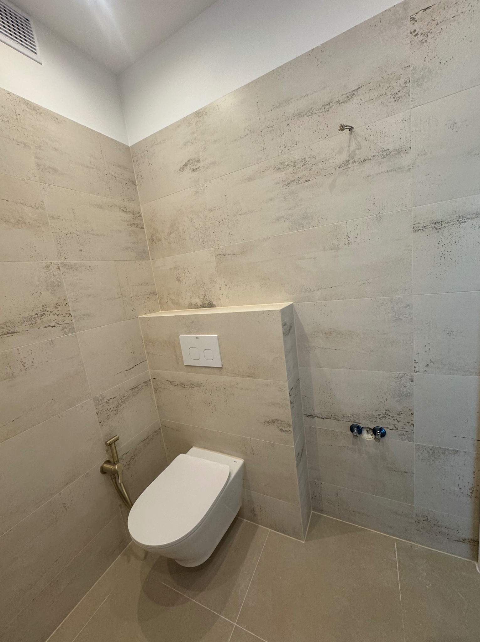 Nowoczesna toaleta podwieszana z bidetką w łazience wykończonej beżowymi płytkami imitującymi trawertyn, widoczne przyłącza wodne i przycisk spłuczki.