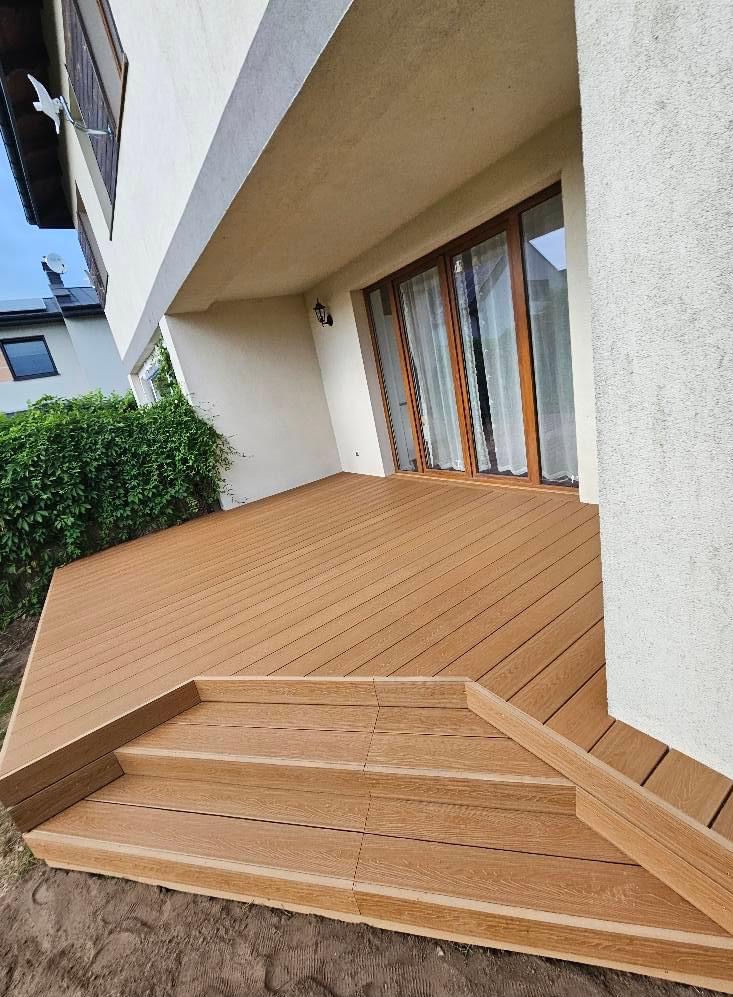 Nowoczesny taras z desek kompozytowych w kolorze jasnego drewna, z trzema schodkami prowadzącymi do drzwi balkonowych, widok z dołu na budynek z jasną elewacją.