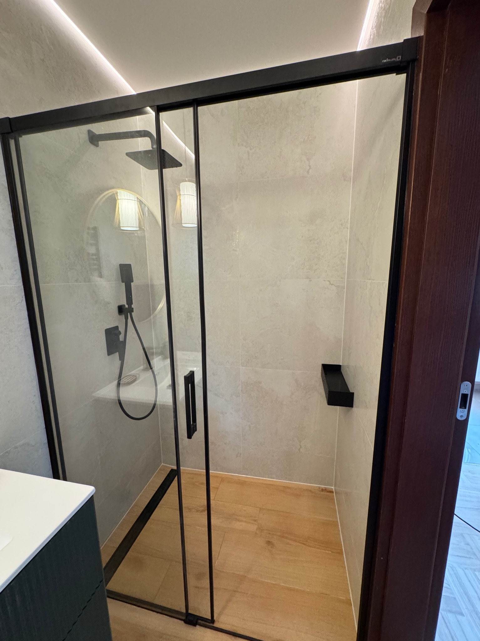 Nowoczesna kabina prysznicowa z czarnymi ramami i deszczownicą, jasne płytki na ścianach, drewnopodobna podłoga, okrągłe lustro z oświetleniem w tle.