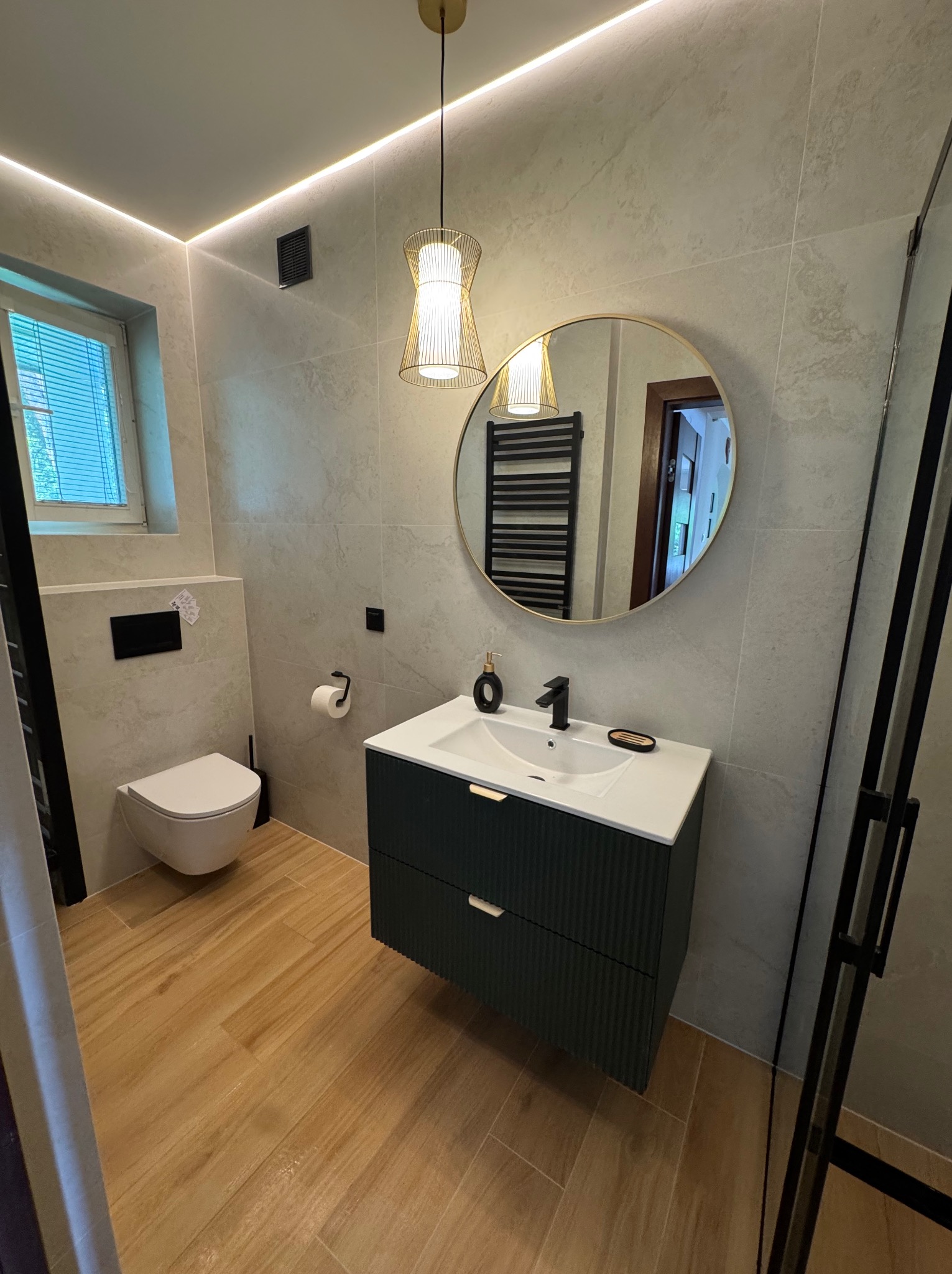 Stylowa łazienka z beżowymi płytkami, drewnopodobną podłogą, umywalką z szafką w kolorze butelkowej zieleni i okrągłym lustrem z elementami złota.