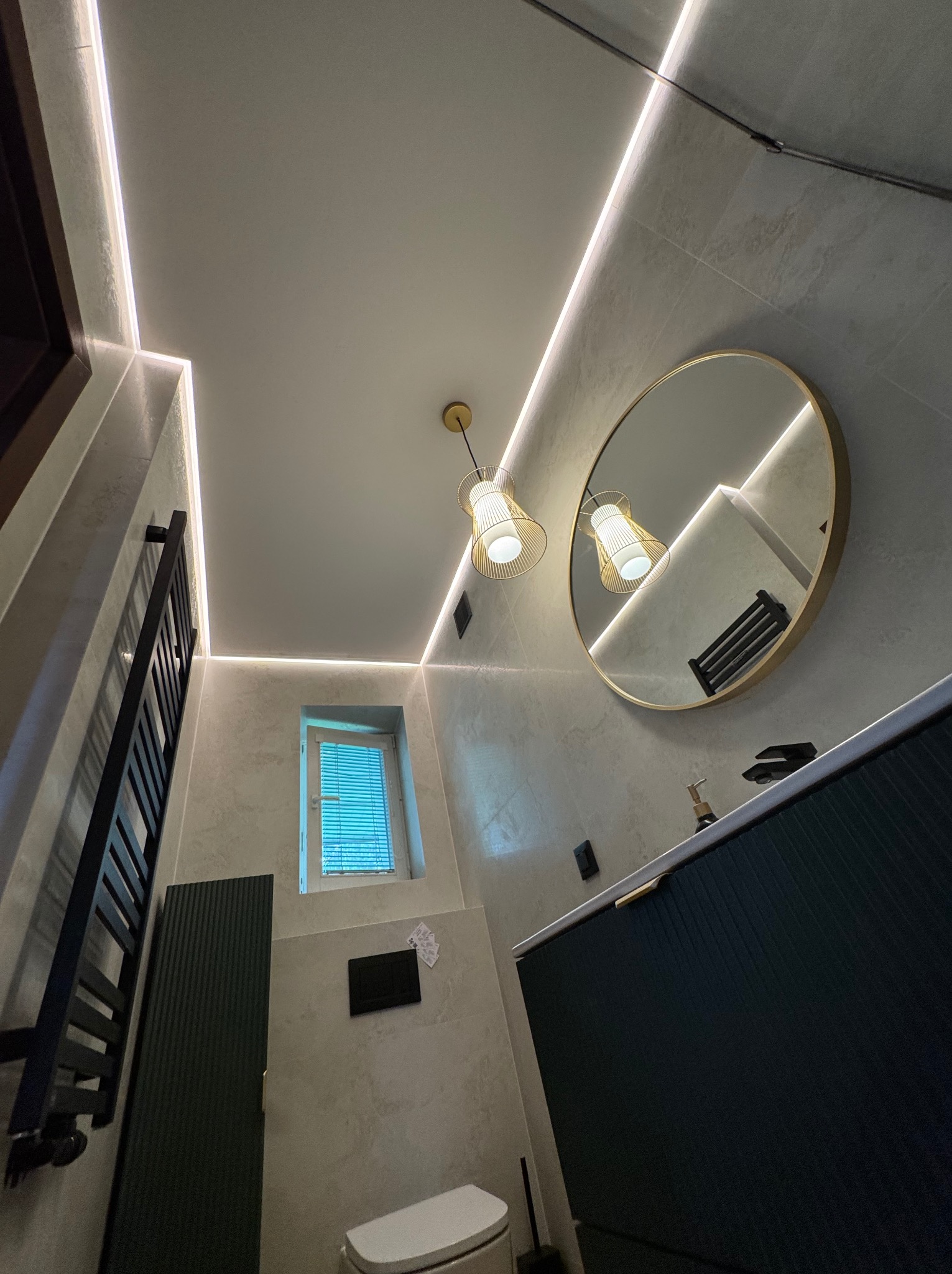 Wnętrze łazienki po remoncie: szare płytki, podświetlane sufity, okrągłe lustro z złotą ramą, czarne meble łazienkowe i designerskie lampy.