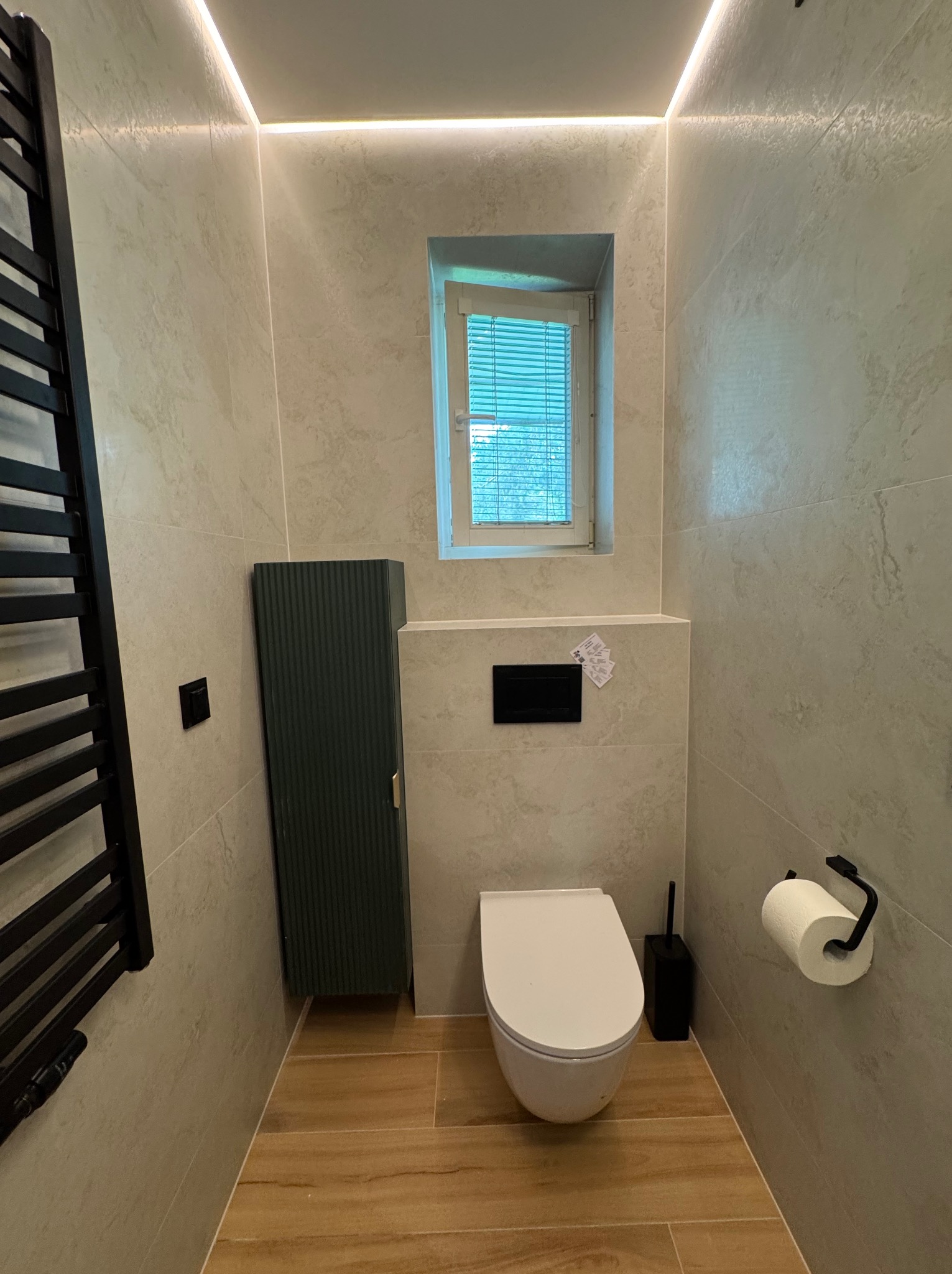 Nowoczesna łazienka z beżowymi płytkami, drewnopodobną podłogą i oświetleniem LED. Uwagę zwraca wisząca toaleta i designerski grzejnik drabinkowy w czarnym kolorze.