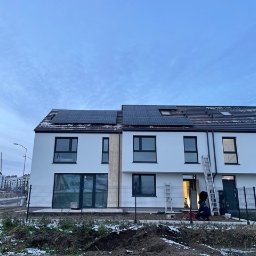 STANDARD ELECTRO SPÓŁKA Z OGRANICZONĄ ODPOWIEDZIALNOŚCIĄ - Montaż paneli słonecznych na dachu szeregowca, widoczna drabina i pracownik w kombinezonie, w tle zabudowa wielorodzinna i pochmurne niebo.