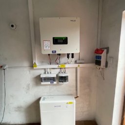 STANDARD ELECTRO SPÓŁKA Z OGRANICZONĄ ODPOWIEDZIALNOŚCIĄ - Wewnętrzna instalacja: falownik Sofar z zielonym wyświetlaczem, obok licznik energii i biały magazyn energii Sofar na tle szarej ściany z widocznymi śladami po instalacji.