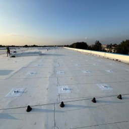 STANDARD ELECTRO SPÓŁKA Z OGRANICZONĄ ODPOWIEDZIALNOŚCIĄ - Biały dach przemysłowy z elementami montażowymi fotowoltaiki i świetlikami dachowymi, w tle zabudowa jednorodzinna, widok z góry.