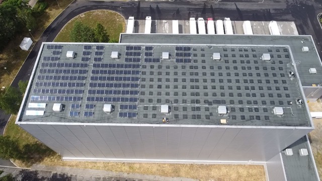 Widok z góry na dach dużego budynku przemysłowego z częściowo zainstalowanymi panelami fotowoltaicznymi; w tle zaparkowane białe ciężarówki.