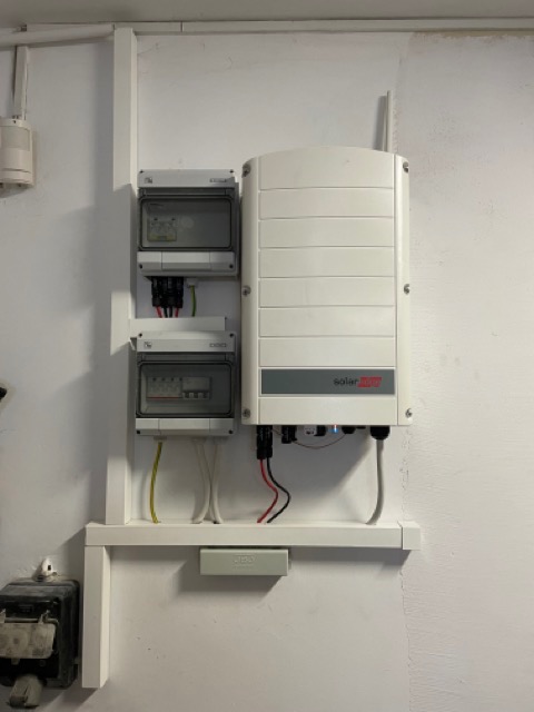 Biały falownik SolarEdge zamontowany na ścianie obok dwóch rozdzielnic elektrycznych w białej obudowie.