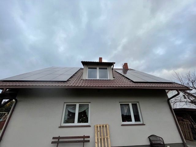 Dom jednorodzinny z brązowym dachem pokrytym panelami słonecznymi, widok z dołu na tle pochmurnego nieba.