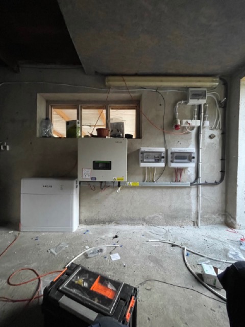 Wnętrze pomieszczenia z zamontowanym falownikiem fotowoltaicznym, akumulatorem energii, rozdzielnicami elektrycznymi i przewodami, widoczne ślady prac instalacyjnych na podłodze, narzędzia w skrzynce.