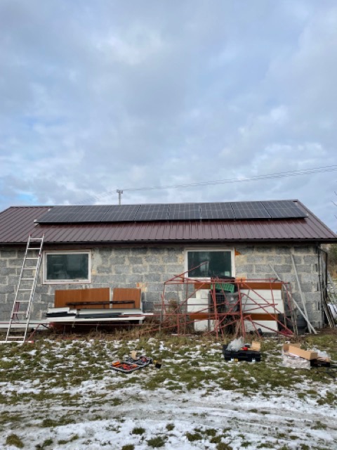 Szary budynek z brązowym dachem, na którym zamontowano panele słoneczne; na ziemi leżą narzędzia i częściowo zalega śnieg. Przystawiona drabina do ściany budynku.