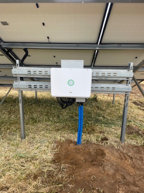 Widok od dołu na moduły fotowoltaiczne z białym inwerterem z zielonym okręgiem na obudowie, zamontowanym na metalowej konstrukcji, z niebieskimi rurami prowadzącymi do ziemi.