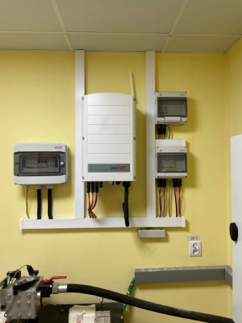 Biały falownik solarny marki SolarEdge zamontowany na żółtej ścianie wewnątrz pomieszczenia, widoczne również skrzynki elektryczne i fragment instalacji hydraulicznej.