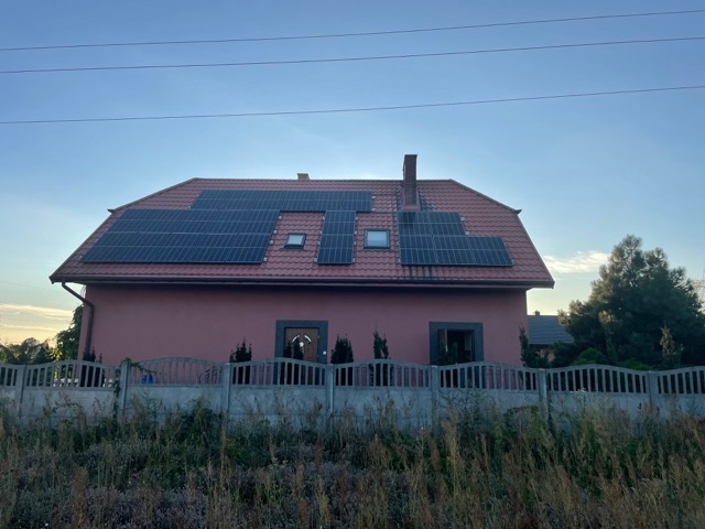 Dom jednorodzinny z zainstalowanymi panelami słonecznymi na dachu pokrytym czerwoną dachówką, widok zza betonowego ogrodzenia porośniętego roślinnością.