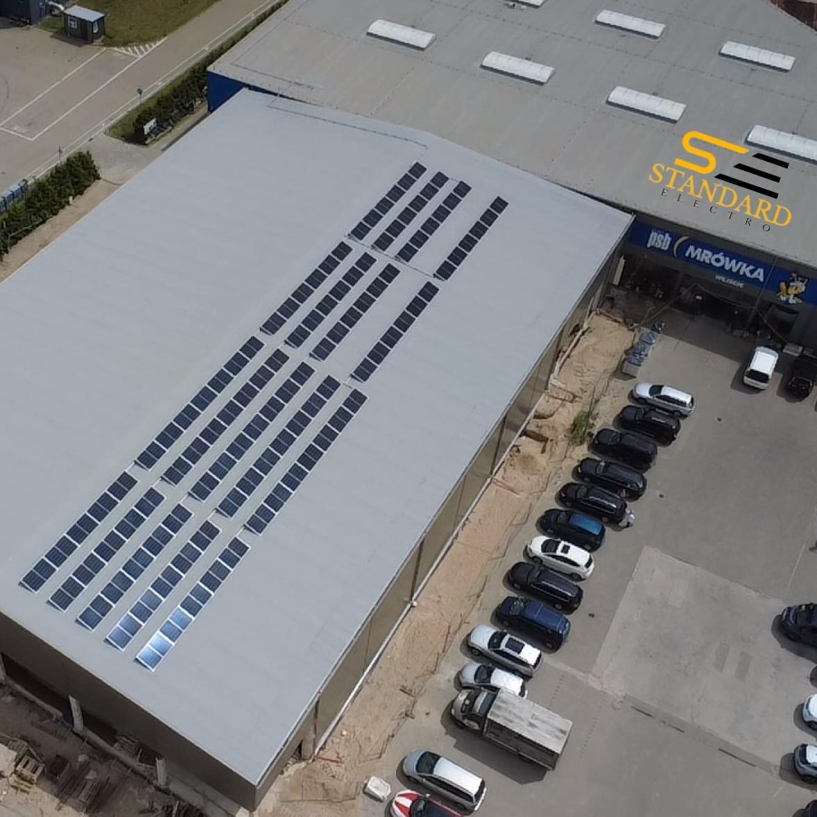 Fotowoltaika na dachu budynku handlowego z parkingiem. Panele ułożone w rzędach, widok z góry. Logo firmy na budynku.