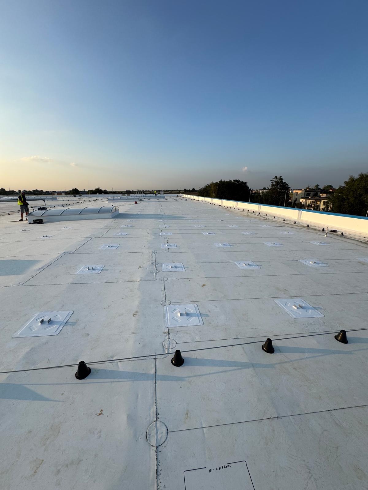 Biały dach przemysłowy z elementami montażowymi fotowoltaiki i świetlikami dachowymi, w tle zabudowa jednorodzinna, widok z góry.