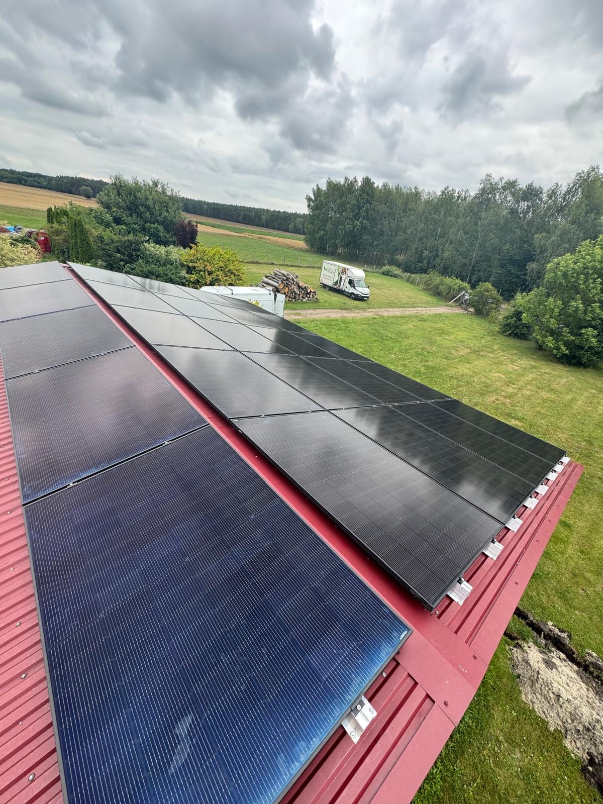 Panele fotowoltaiczne na czerwonym dachu z blachy, widok z góry na tle zieleni i pochmurnego nieba. W oddali zaparkowany biały bus.