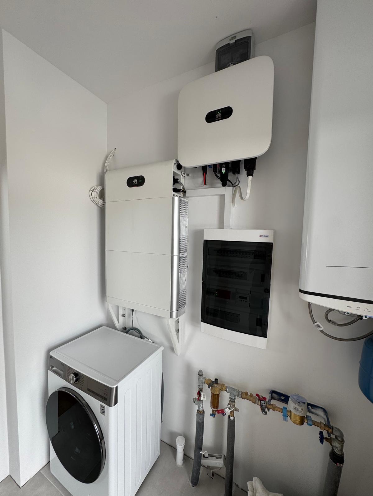Białe urządzenia fotowoltaiczne Huawei zamontowane na ścianie w pomieszczeniu gospodarczym obok pralki i rozdzielnicy elektrycznej. Widoczne rury i instalacje.