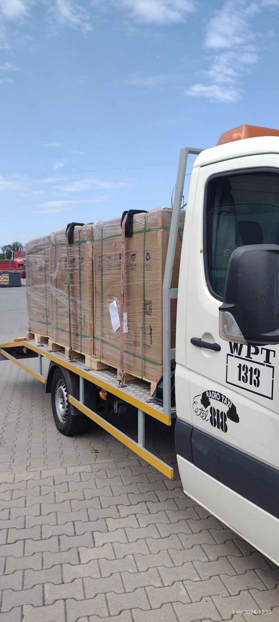 Biała laweta z rampą załadowaną paletami z kartonowymi paczkami, zabezpieczonymi folią i pasami transportowymi, widoczne logo firmy przewozowej na drzwiach.