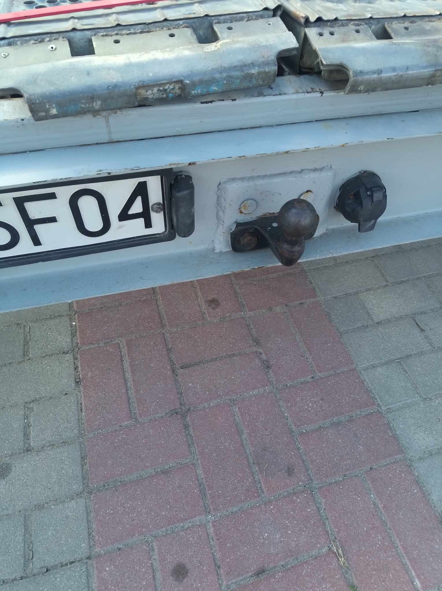 Hak holowniczy z gniazdem elektrycznym i tablicą rejestracyjną przyczepy na tle kostki brukowej.