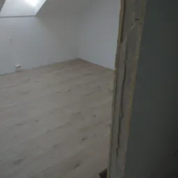 Jasne panele podłogowe ułożone w pokoju na poddaszu z oknem dachowym, widoczny fragment ościeżnicy z surowym wykończeniem.