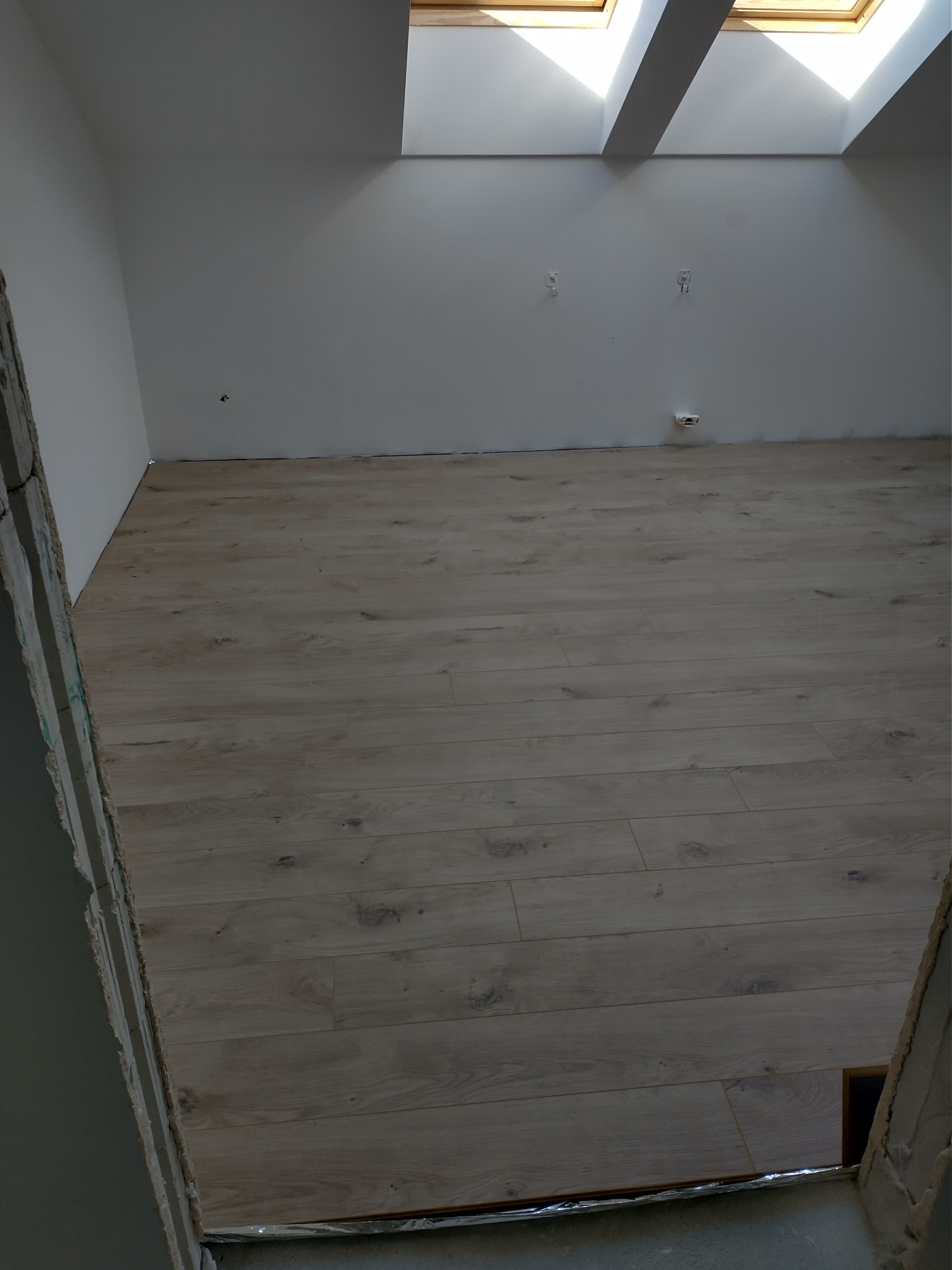 Jasne panele podłogowe ułożone w pokoju z oknami dachowymi, widok zza surowej, nieotynkowanej krawędzi ściany.