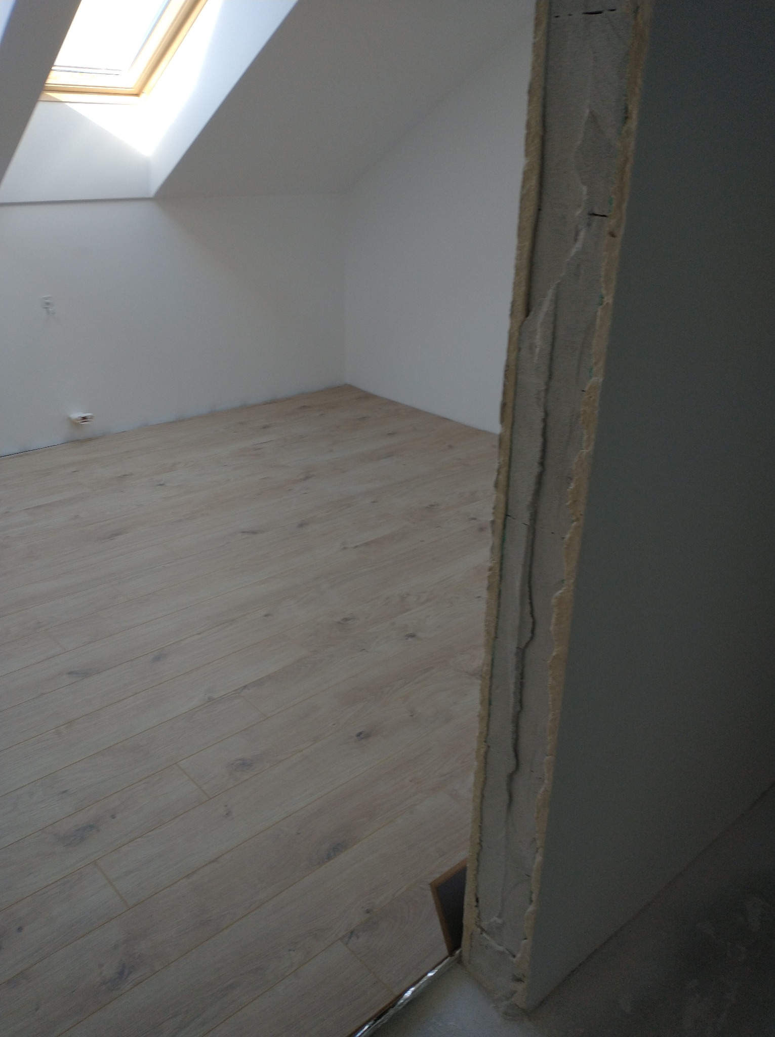 Jasne panele podłogowe ułożone w pokoju na poddaszu z oknem dachowym, widoczny fragment ościeżnicy z surowym wykończeniem.