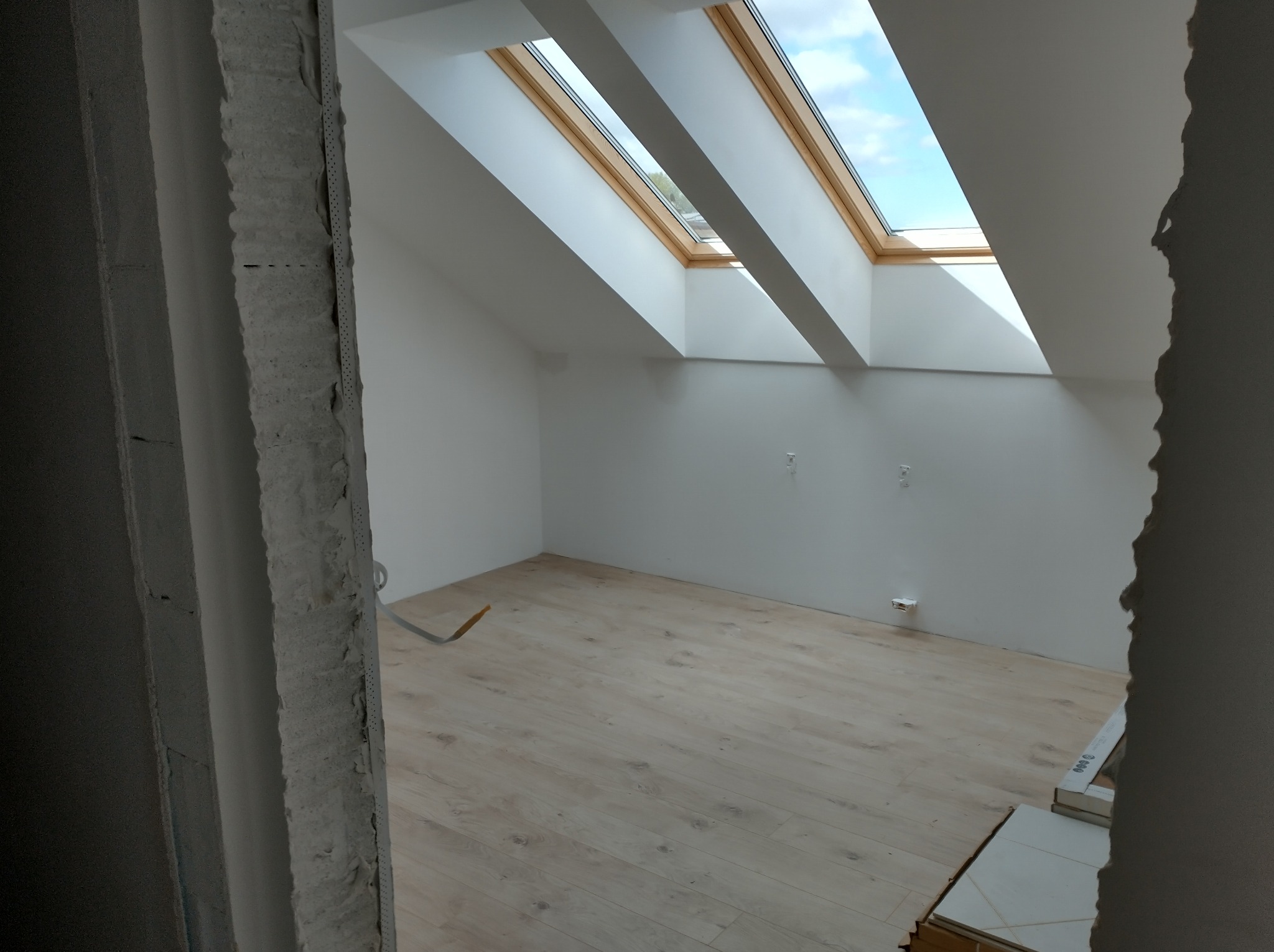 Poddasze z trzema oknami dachowymi, świeżo ułożone panele podłogowe w odcieniach beżu, ściany pomalowane na biało, widoczne puszki instalacyjne, surowy ościeżnica po lewej stronie.