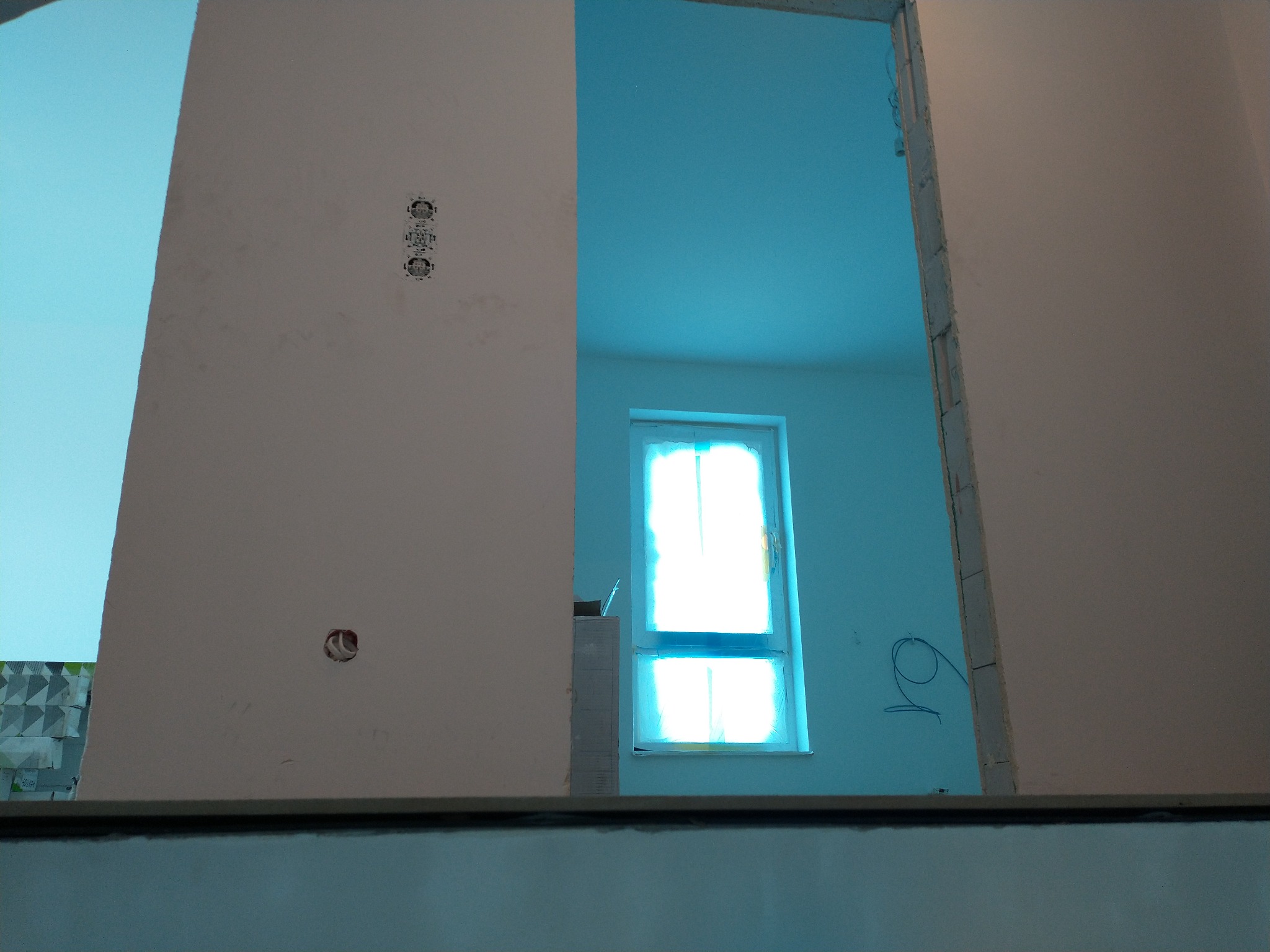Wnętrze w trakcie wykańczania, widoczne ściany z gniazdkami elektrycznymi, surowe obramowanie otworu drzwiowego, okno z zasłoniętą szybą i niebieskie ściany w sąsiednim pomieszczeniu.