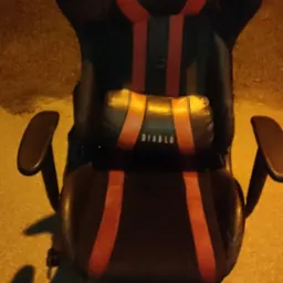 Czarne krzesło gamingowe z czerwonymi akcentami i poduszką lędźwiową z napisem 'DIABLO' na szorstkim, oświetlonym podłożu.