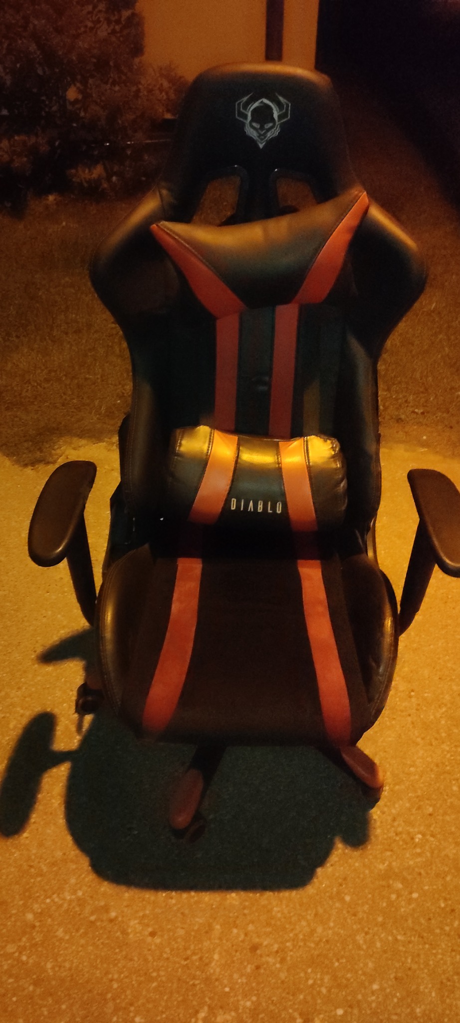 Czarne krzesło gamingowe z czerwonymi akcentami i poduszką lędźwiową z napisem 'DIABLO' na szorstkim, oświetlonym podłożu.