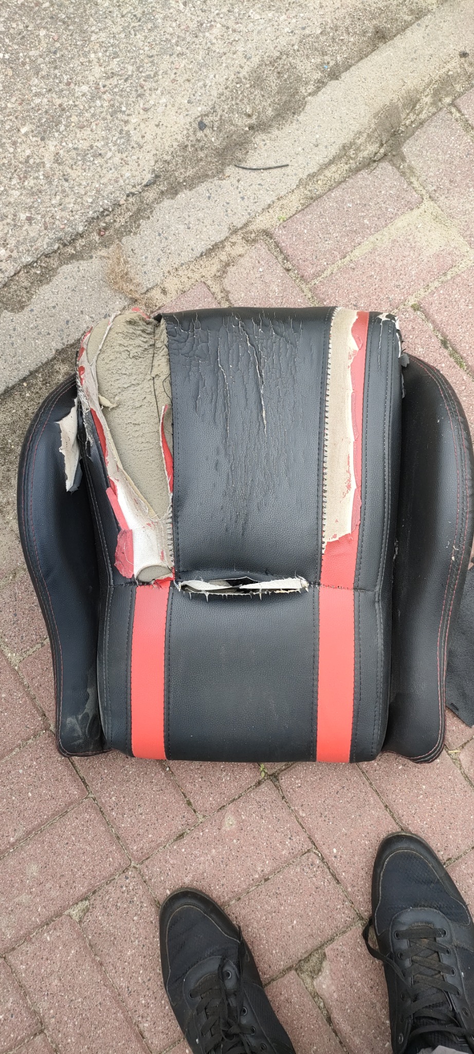 Zniszczone, podarte krzesło gamingowe z czarno-czerwonej skóry ekologicznej leżące na chodniku z kostki brukowej, widoczne fragmenty gąbki i podszewki, obok stopy w czarnych butach sportowych.