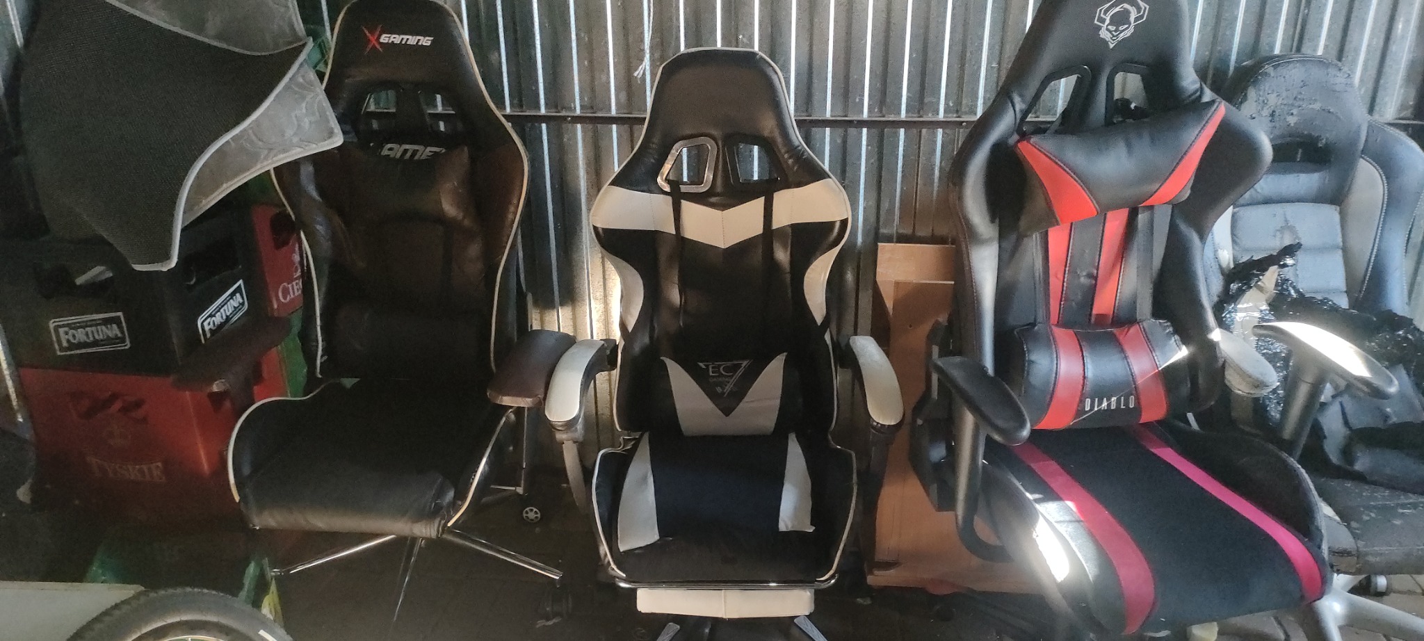 Trzy fotele gamingowe różnych marek (X-Gaming, EC, Diablo) ustawione obok siebie na tle blaszanej ściany, w pomieszczeniu o słabym oświetleniu, z widocznymi elementami otoczenia, w tym skrzynkami...