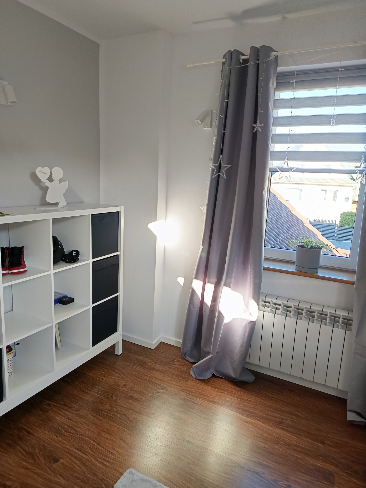 Jasne wnętrze pokoju z drewnopodobną podłogą, białym regałem z czarnymi wkładami, szarymi zasłonami z gwiazdkami i grzejnikiem pod oknem. Na regale dekoracja z aniołem. Promienie słoneczne wpadają...