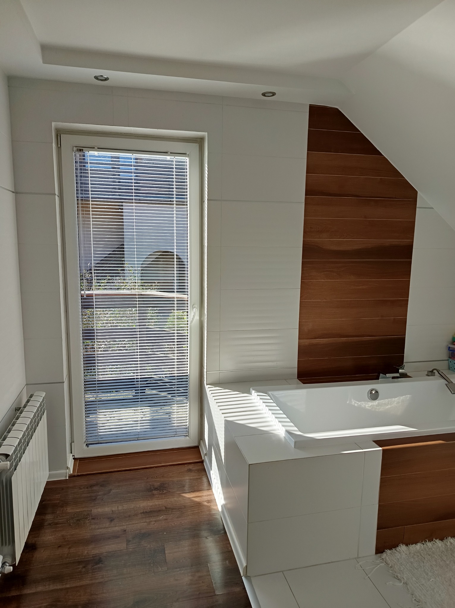 Nowoczesna łazienka na poddaszu z białą wanną obudowaną płytkami i drewnianym panelem, okno z żaluzjami i drewniana podłoga.