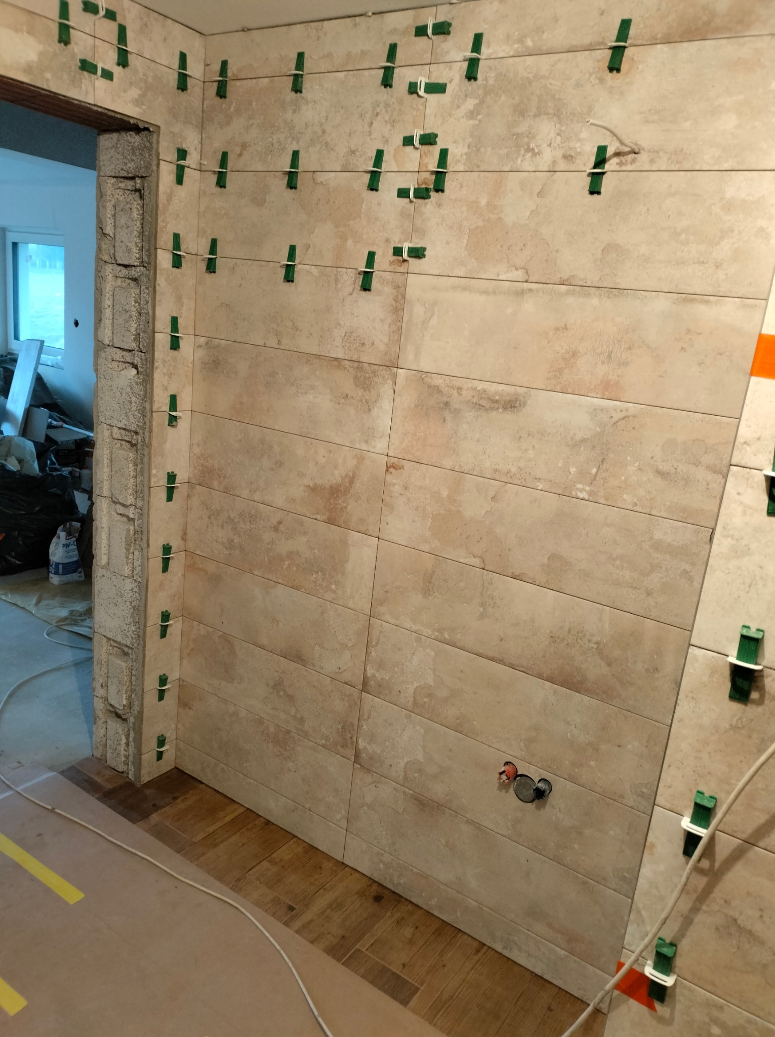 Ściana wyłożona beżowymi, poziomymi płytkami ceramicznymi, z widocznymi zielonymi klipsami poziomującymi i białymi gumkami, fragment podłogi wyłożony płytkami imitującymi drewno, widoczne puszki...