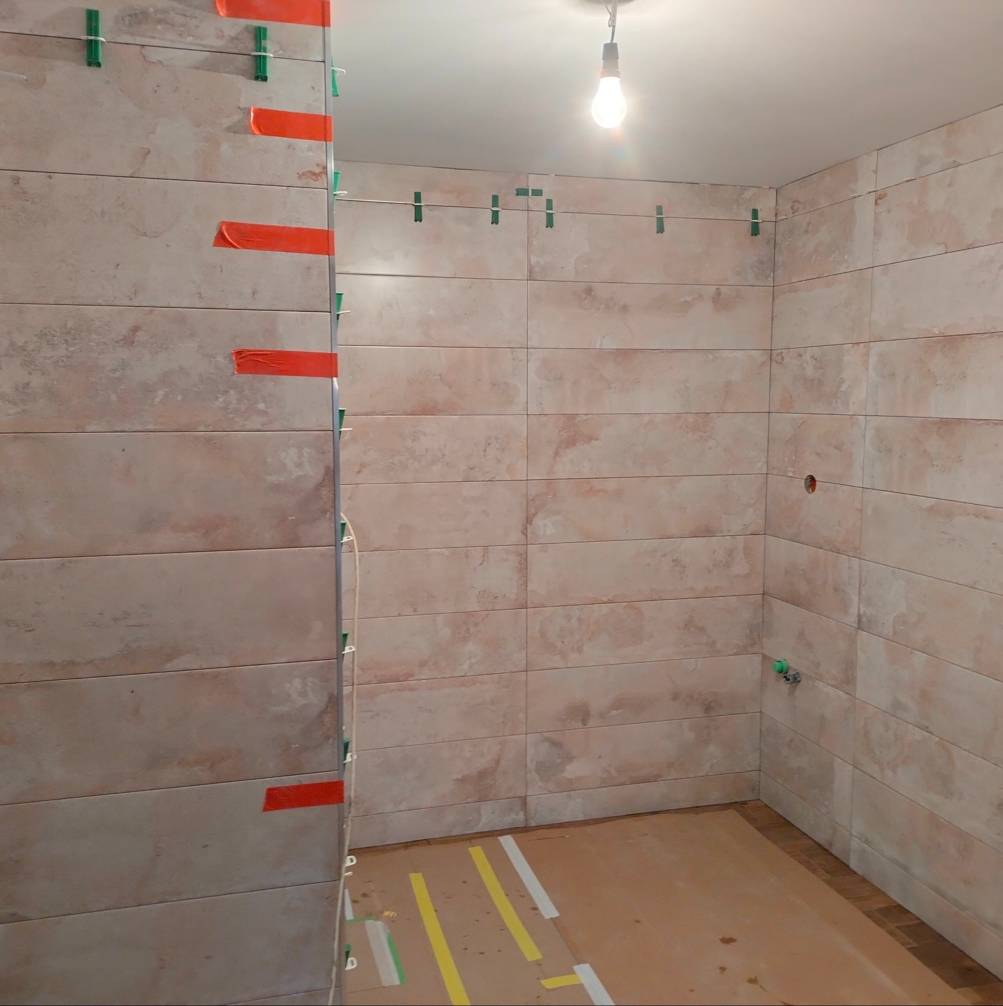 Układanie beżowych, poziomych płytek w łazience z użyciem klipsów poziomujących i taśmy malarskiej, widoczny fragment podłogi zabezpieczony kartonem z taśmą.