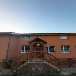 Mariola Starzec - Dwupiętrowy budynek z drewnianym zadaszeniem nad wejściem i barierkami schodów w stylu rustykalnym. Pomarańczowa fasada, białe okna, wieczorne światło.