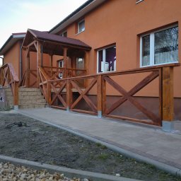 StolMar - Zadaszone wejście do domu z drewnianą balustradą w rustykalnym stylu, z krzyżowymi wzorami. Widok z poziomu chodnika, jesienne popołudnie.