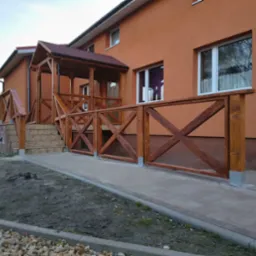 Zadaszone wejście do domu z drewnianą balustradą w rustykalnym stylu, z krzyżowymi wzorami. Widok z poziomu chodnika, jesienne popołudnie.